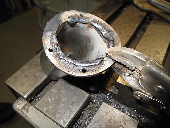 FinishingSteamPipeShroud27-May09 006 With the flange hole to the proper size, it is outside for some welding. Using a temporary table setup, and dodging the raindrops (not a good mix with an AC stick welder) I manage to get the 20 year old 6013 rod to light and put down some beads.  It doesn't look pretty with all the flux on it now, but after some cleaning and grinding back to the curved profile, the shroud is now one piece.
