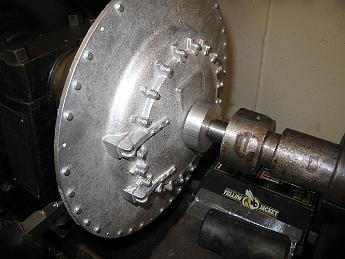 SmokeboxStarted4Feb09 001 4-Feb-09 We start a new section - the Smokebox!  It's always fun to start a new one--fresh drawings to look at, new castings to puzzle out how to hold.  We put the big Rahn-Larmon lathe to use to turn the OD of the smokebox front true, and clean up the back.  Although we have the casting clamped from behind, we use the live center and a spacer block for additional pressure to keep it in place.
